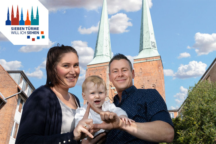 Herr und Frau Kosakowski halten ihr Kleinkind zwischen sich und zeigen mit den Fingern einen Turm | Im Hintergrund sind die Türme des Doms zu Lübeck zu sehen, sowie der blaue Himmel mit ein paar Wolken - Copyright: Rainer Jensen